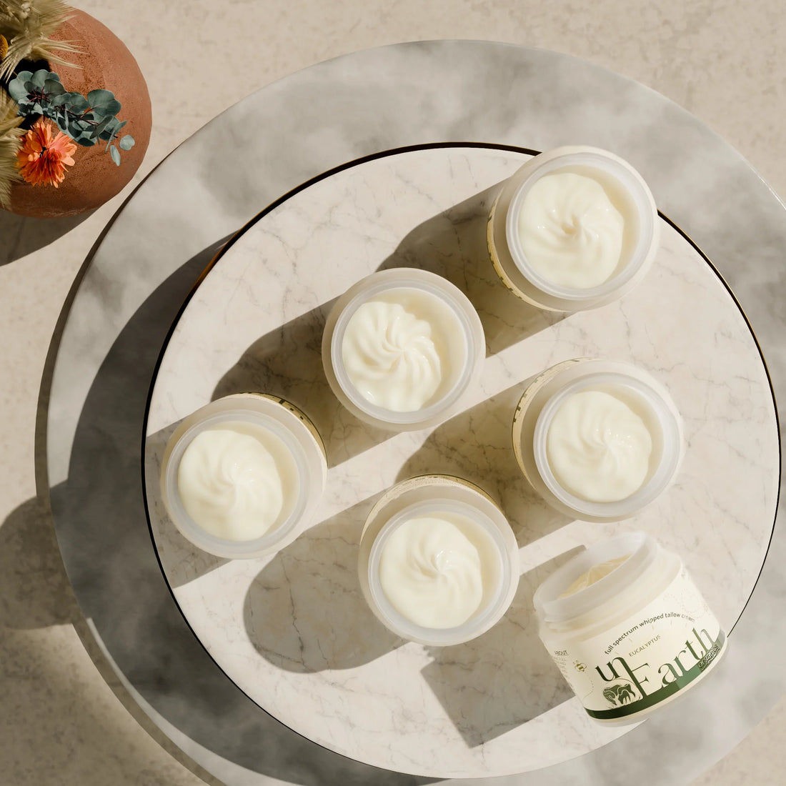 Jars of unscented and eucalyptus whipped tallow moisturizer from unearth organics laying on a flat cream surface.
